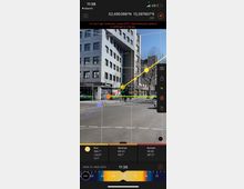 Eine Straßenkreuzung in einer städtischen Umgebung mit einem modernen mehrstöckigen Gebäude im Hintergrund, das eine Fassade aus Stein und Glas hat. Im Vordergrund sind Fahrbahnmarkierungen sowie Fahrräder und ein Fußgängerweg zu sehen. Angaben zu Sonnenstand und Himmelsrichtungen sind digital im Bild eingeblendet.