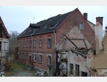 Backstein-Gebäude mit beschädigtem Dach und zerfallener Fassade, daneben Ruinen eines einstigen Gebäudes mit eingestürzten Wänden und freiliegendem Holzwerk. Im Hintergrund ist ein bewachsener Außenbereich zu erkennen.