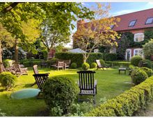 Gepflegter Garten mit grünem Rasen, umgeben von niedrigen Buchsbaumhecken. Im Vordergrund stehen mehrere Holzstühle und -tische, im Hintergrund ist ein Backsteingebäude mit roten Dachschindeln und begrünten Fassaden zu sehen.