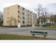 Mehrfamilienhaus mit vier Stockwerken in beige-grauer Fassade und rosa Balkonen, umgeben von kahlen Bäumen. Im Vordergrund befindet sich eine grüne Parkbank neben einer Straße, auf der mehrere Fahrzeuge geparkt sind.