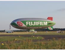 Ein großer grün-weißer Zeppelin mit der Aufschrift „Fujifilm“ steht auf einer Freifläche nahe einer Start- oder Landebahn. Im Vordergrund sind Grasflächen mit gelben Markierungen, im Hintergrund stehen leichte Baumreihen und Menschen in weißer Kleidung.