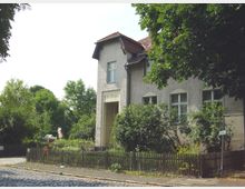 Eine zweistöckige Schule im traditionellen Baustil mit heller Putzfassade und roten Dachziegeln, umgeben von Bäumen und einem Holzzaun im Vorgarten. Der Schriftzug „Schulhaus“ ist über dem Eingang zu sehen, und die Umgebung zeigt eine gepflasterte Straße und begrünte Flächen.