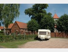 Ein Wohnwagen steht auf einem sandigen Platz vor einem Backsteinhaus mit rotem Ziegeldach und einer Hecke. Im Hintergrund sind Bäume sowie der Turm eines Fachwerkgebäudes mit rotem Spitzdach sichtbar.