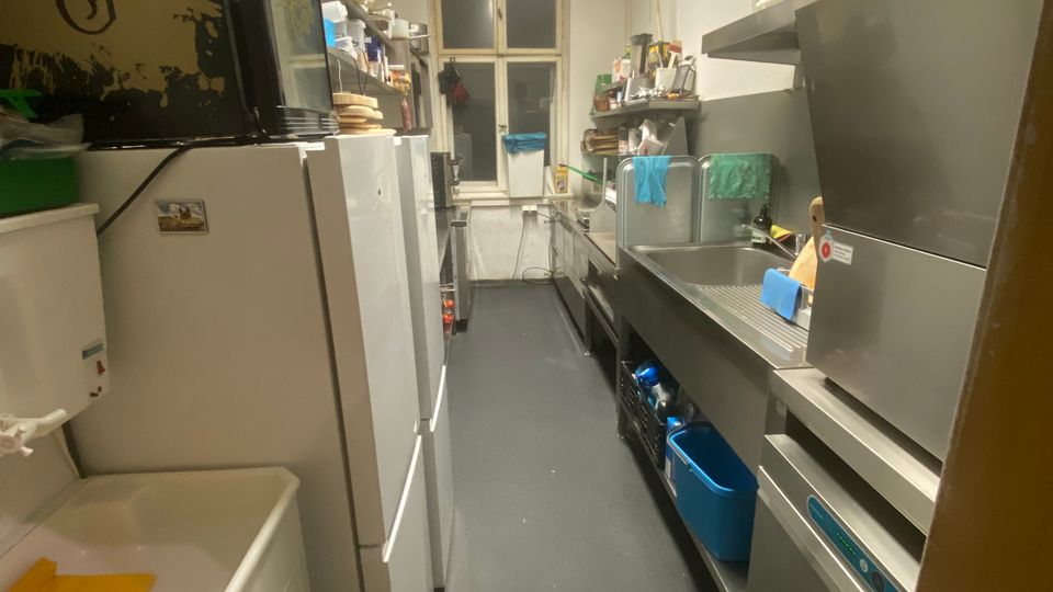 Schmaler, länglicher Küchen- und Spülraum mit grauem Boden, Deckenleuchten und einem Fenster am Ende. Links stehen Kühlschränke und Regale, rechts verläuft eine Edelstahl-Arbeitszeile mit Spülbecken, Abtropffläche und großen Küchengeräten.