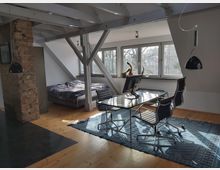 Helles Dachgeschosszimmer mit einem Arbeitsbereich und Schlafbereich. Sichtbare Holzbalken, eine freiliegende Backsteinwand, ein Glastisch mit Bürostühlen auf einem blauen Teppich, ein Bett in der Ecke und große Fenster mit Blick auf Bäume.