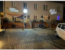 Außenansicht eines Gebäudes bei Nacht mit neonbeleuchteten Schildern "Tattoo" und "Road House". Vor dem Gebäude befinden sich ein gepflasterter Platz, eine parkende weiße Limousine und ein Motorrad neben einer Treppe, die zum Eingang hinaufführt.