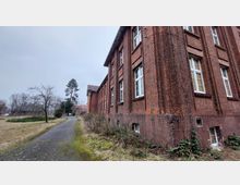 Backsteinbau mit weißen, rechteckigen Fenstern, umgeben von einer Wiese und kahlen Bäumen. Ein schmaler Gehweg führt an der Gebäudeseite entlang, ergänzt durch spärliche Vegetation und einen trüben Himmel im Hintergrund.