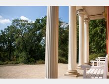 Säulengang mit weißen, kannelierten Säulen und verzierten Kapitellen, daneben weiße Sitzbänke; im Hintergrund ein sandiger Boden und ein parkähnliches Gelände mit Bäumen unter klarem Himmel.