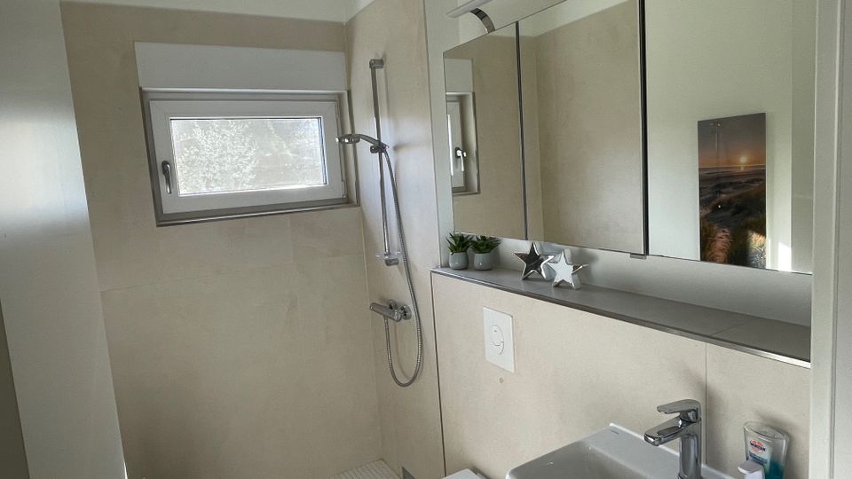 Modernes Badezimmer mit beiger Wandverkleidung, einem kleinen Fenster, einer Dusche mit Handbrause und einer weißen Toilette. Ein Waschbecken mit Spiegel und Ablage, auf der Dekorationen und Pflanzen stehen, befindet sich rechts.