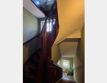 Innenansicht eines schmalen Treppenhauses mit dunkel gebeizter Holztreppe und geschwungenem Geländer, das über mehrere Etagen verläuft. Helle Fenster beleuchten die grünlich gestrichenen Wände, unten steht eine Topfpflanze am Treppenabsatz.