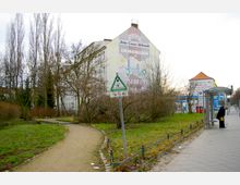 Parkähnliche Grünfläche mit einem geschwungenen Kiesweg, umgeben von kahlen Bäumen und Sträuchern im Winter. Im Hintergrund ein großes Wohngebäude mit Werbung auf der Fassade sowie eine Bushaltestelle mit einer wartenden Person.