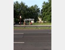 Bushaltestelle an einer Straße mit mehreren Fahrspuren, umgeben von Grünflächen und großen Bäumen im Hintergrund. Einige Menschen warten unter der Überdachung der Haltestelle.