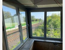 Blick durch ein großes, mehrteiliges Fenster auf Bahngleise, die von grüner Vegetation und einem Lärmschutzzaun aus Metall begleitet werden. Im Vordergrund ist ein Teil eines Innenraums mit unverputzter Wand sichtbar.