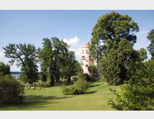 Schloss inmitten einer weitläufigen Parklandschaft mit grüner Wiese, hohen Bäumen und vereinzelten Büschen. Im Hintergrund ist ein Fluss mit einer Brücke zu sehen, der Himmel ist blau mit einigen Wolken.
