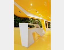 Modernes Innenraumdesign mit gelben Böden und Decken, verziert mit weißen Illustrationen; eine weiße Empfangstheke mit kantigem Design steht im Vordergrund. Eine grüne Wand mit vertikaler Bepflanzung bildet einen Kontrast zur leuchtenden Farbgestaltung.