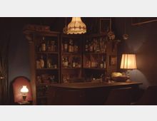 Ein gemütlicher Barbereich mit schummriger Beleuchtung, ausgestattet mit einem Holzregal voller Flaschen und Gläser, einer Theke mit Stühlen und einer Tischlampe mit warmem Licht. Im Hintergrund hängen ein Kronleuchter und eine Tafel mit Schriftzügen.