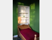 Holzbettrahmen mit roter Bettdecke in einem Raum mit grünen Wänden; ein kleines Regal mit einer Lampe und einem grünen Glas steht in einer Wandnische. Lichteinfall durch ein Fenster wirft Schatten auf die Wände.