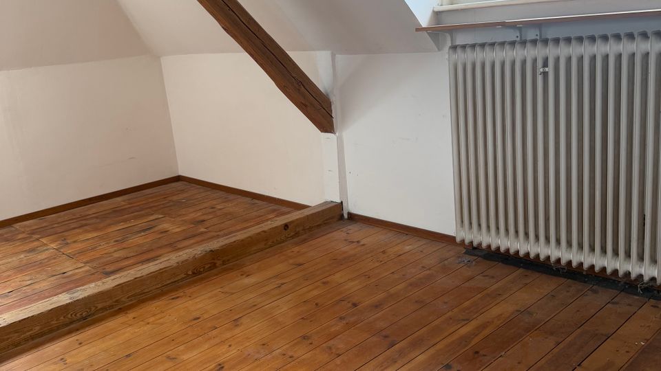 Leerer Dachgeschossraum mit schrägen weißen Wänden, sichtbarem Holzbalken und breiten Holzdielenboden mit kleiner Stufe. Rechts ein Fenster mit Blick ins Grüne über einem großen Rippenheizkörper.