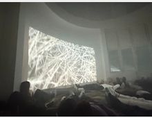 Ein abgedunkelter Raum mit Menschen, die auf Matten am Boden liegen und auf eine große Wandprojektion blicken, die helle, verwobene Linien zeigt. Im Hintergrund befinden sich hohe Fenster, die teilweise verhangen sind.