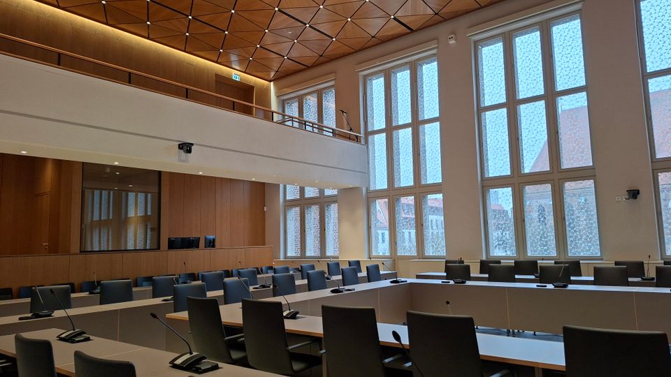 Moderner Sitzungssaal mit U-förmig angeordneten Tischen, schwarzen Stühlen und integrierten Mikrofonen. Hohe, mehrteilige Fensterfronten und eine hölzerne Wand- und Deckenverkleidung mit Galerieebene prägen den Raum.