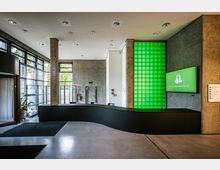 Moderner Empfangsbereich mit gebogener schwarzer Rezeption, grün beleuchteter Glaswand und Bildschirm mit der Aufschrift „Besondere Orte“. Große Fensterfronten lassen Tageslicht herein, während Betonwände und ein heller Boden die Umgebung prägen.