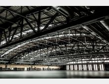 Große, leere Industriehalle mit sichtbarer Metallkonstruktion und Stahlträgern an der Decke. Die Wände am Rand sind mit hohen Fenstern versehen, durch die diffuses Licht einfällt.