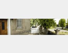 Panorama eines Eingangsbereichs eines Gebäudes mit heller Wand, einem Fenster mit Rollladen und einer Holztür links. Im Vordergrund ein gepflasterter Boden, rechts ein Baum und im Hintergrund ein kleiner Zaun sowie eine kurvige, von Bäumen gesäumte Straße.