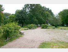 Ein Park mit gepflastertem Weg, umgeben von Grünflächen, Bäumen und Büschen. Mehrere Steinbänke säumen den Weg, der sich durch die gepflegte Landschaft schlängelt.
