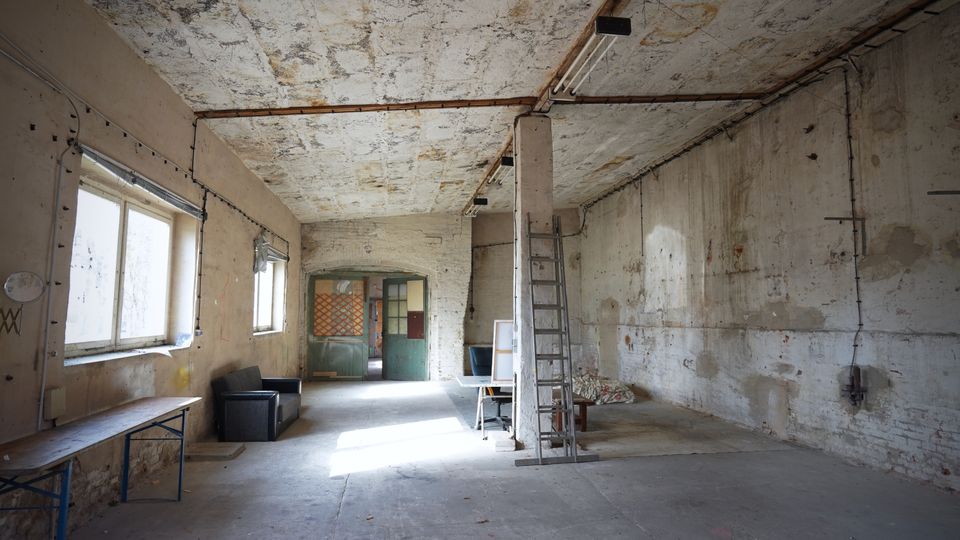 Großer, leerer Innenraum einer alten Industrie- oder Lagerhalle mit rauen, teils abblätternden Wänden und Decke sowie freiliegenden Leitungen und Neonröhren. Links sind mehrere Fenster und eine lange Werkbank, in der Mitte eine Stütze mit angelehnter Leiter, hinten eine grüne Doppeltür und vereinzelte Möbel.