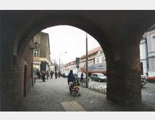 Blick durch einen gemauerten Torbogen auf eine gepflasterte Straße mit Fußgängern, geparkten Autos und Reihen von historischen Wohnhäusern mit Satteldächern im Hintergrund. Links ist ein Geschäftseingang mit einer Laterne sichtbar.