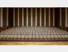 Innenansicht eines Hörsaals oder Auditoriums mit mehreren Reihen gepolsterter Sitze auf ansteigenden Ebenen. Im Hintergrund befinden sich hohe weiße Säulen, die von braunen Wandpaneelen und fensterartigen Elementen mit Gittermuster eingerahmt sind.