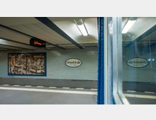U-Bahn-Station Uhlandstraße in Berlin mit hellgrauen Fliesen an den Wänden, einem ovalen Stationsschild und einem Wandbild, das historische Straßenbahnen zeigt. Im Vordergrund ist der Blick durch das Fenster einer Bahn erkennbar.