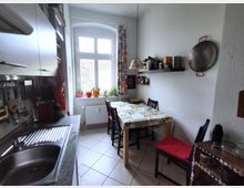 Narrow galley kitchen with a stainless sink and countertop on one side and a small dining table with chairs on the other. A tall arched window with patterned curtains and potted plants brings in light, with wall shelves, hanging cookware, and white tile flooring.