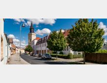 Straßenszene in einer kleinen Stadt mit Kopfsteinpflaster, Bäumen und geparkten Autos auf der rechten Straßenseite. Im Hintergrund steht ein Kirchturm mit dunkler Spitzhaube und umliegenden Gebäuden mit roten Ziegeldächern.