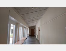 Ein heller Flur in einem Gebäude mit weiß gestrichenen Wänden, rechteckigen Deckenleuchten und bodentiefen Fenstern auf der linken Seite. An der rechten Wand befinden sich Türen und ein Feuerlöscher, während mehrere schmale Tische entlang der Fenster aufgereiht sind.