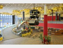 Innenansicht eines öffentlichen Gebäudes mit großer Halle, umfasst von breiten Treppen und einer Rampe. Die Halle verfügt über eine Spiel- und Kletterstruktur mit Rutschen, hängende schwarze Lautsprecher, rote Vorhänge und Sitzbereiche mit Tischen und Stühlen; die Decke ist mit geometrischen, gelben Deckenelementen gestaltet.