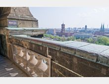 Blick von einer historischen Balkonplattform mit grünlich oxidierten Kupferelementen auf die Stadt Berlin, darunter das Rote Rathaus und Kirchtürme sowie grüne Parkanlagen. Ein kleines Schild mit "Ausgang" ist an der Wand angebracht.
