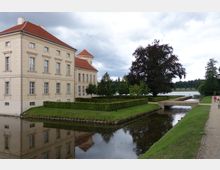Schlossgebäude mit beigem Putz und roten Ziegeldächern, umgeben von gepflegten Grünanlagen und einem Wassergraben. Eine kleine Brücke führt über den Graben, im Hintergrund sind ein Baum, Spazierwege und ein See zu sehen.
