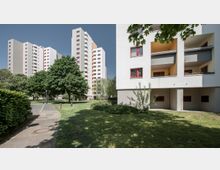 Wohnsiedlung mit mehreren mehrstöckigen Wohngebäuden in moderner Architektur, umgeben von Grünflächen und Bäumen. Im Vordergrund ein Gehweg, Wiesenflächen und ein niedriger Gebäudeteil mit Balkonen.