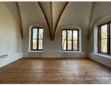 Ein leerer Raum mit Holzfußboden, einer gewölbten Decke mit Holzbalken und drei hohen, bogenförmigen Fenstern, die durch Gitter und Vorhänge gerahmt sind. Außen sind Bäume und ein Gebäude sichtbar.