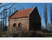 Backsteinbau mit Satteldach und roten Dachziegeln in ländlicher Umgebung; zwei Rundbogenfenster und ein großes Tor prägen die Vorderseite. Im Hintergrund sind kahle Pappeln und blauer Himmel zu sehen.