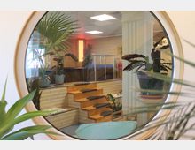 Ein runder Fensterrahmen bietet Einblick in einen Innenraum mit Holztreppen, tropischen Pflanzen und einem Whirlpool. Die warm beleuchtete Umgebung weist Holzvertäfelungen, dekorative Säulen und eine entspannte Atmosphäre auf.