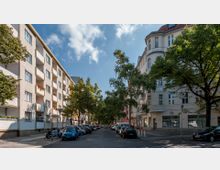 Straßenszene in einer Wohngegend mit mehrstöckigen Gebäuden links und rechts, hellen Fassaden und Balkonen auf der linken Seite, sowie einem Eckgebäude mit abgerundeter Architektur rechts. Die Straße ist von Bäumen gesäumt und beidseitig mit parkenden Autos belegt.