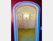 Ein Flur mit grünem Teppichboden, eingerahmt von einer weißen, abgerundeten Türöffnung. Im Hintergrund sind zwei weiße Türen mit goldenen Verzierungen und ein zentral angebrachtes Wandlicht mit zwei Leuchten sichtbar.