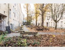 Hinterhof einer Wohnanlage mit verblassten, weiß gestrichenen Fassaden. Im Hof stehen Bäume mit Herbstlaub, verstreuter Müll, Bänke und ein gepflasterter Gehweg, umgeben von Laub und Gestrüpp.