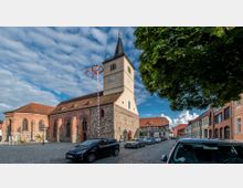 Eine historische Kirche mit einem rechteckigen Turm mit Zifferblättern einer Uhr und einem spitzen Dach, umgeben von Fachwerkhäusern und einer gepflasterten Straße mit parkenden Autos. Rechts im Bild sind Bäume, die Schatten spenden, und links ein geschmückter Maibaum.