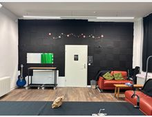 Interior of a small studio lounge with a black acoustic foam wall and a central white door, wood flooring, and bright fluorescent ceiling lights. A red leather sofa with cushions sits beside a small wooden coffee table, with a vanity-style mirror and table on wheels and a guitar along the left wall.