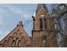 Backsteinkirche im neugotischen Stil mit spitzem Turm und mehreren kleinen Rundbogenfenstern. Im Vordergrund hängen die kahlen Äste eines Baumes, der die Architektur teilweise verdeckt.