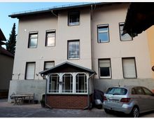 Hinterhofansicht eines mehrstöckigen Wohnhauses mit heller Putzfassade, mehreren rechteckigen Fenstern und einem kleinen verglasten Anbau mit Satteldach und Ziegelsockel. Im gepflasterten Hof stehen ein geparktes Auto, daneben eine Bank mit Tisch sowie einige Pflanzkübel.