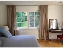 Schlafzimmer mit großem Fenster, das von beigen Vorhängen eingerahmt ist. Der Raum hat Holzboden, ein Bett mit blauer Bettwäsche, einen Heizkörper unter dem Fenster und einen kleinen Tisch mit einem gerahmten Bild sowie weißen Dekovasen darauf. Draußen sind grüne Bäume und ein Garten sichtbar.